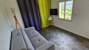 Ett rum med en grå soffa, ett fönster med utsikt över grönområden, en väggmonterad TV och en gardin.