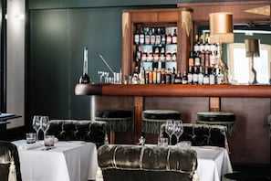 Zona de bar con mostrador de madera, botellas, copas y una lámpara. Hay sillas acolchadas y mesas con copas de vino y cubiertos.