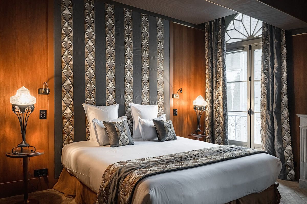 Hotellihuone, jossa on suuri sänky, kaksi yöpöytävalaisinta, ikkuna, jossa on verhot, ja kuviollinen seinä.
