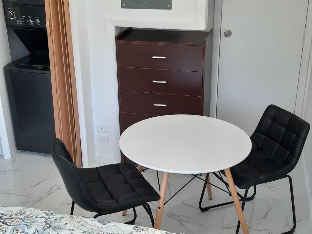 A compact room with a white round table and two black chairs, a dark brown cabinet with drawers, and a black appliance in the background.