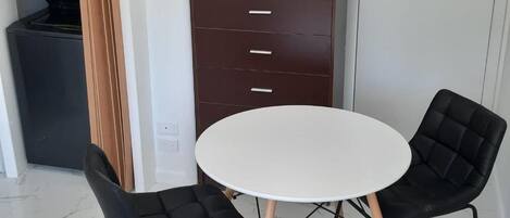 A compact room with a white round table and two black chairs, a dark brown cabinet with drawers, and a black appliance in the background.