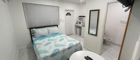 Un dormitorio con cama, mesita de noche, espejo, ventana con persianas, microondas y retrete.