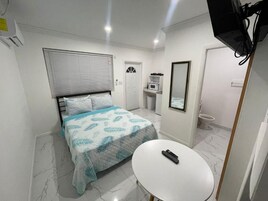 Un dormitorio con cama, mesita de noche, espejo, ventana con persianas, microondas y retrete.