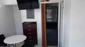 A compact room with a bed, a small table, a TV mounted on the wall, a wooden cabinet, and a built-in oven.