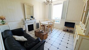 A living room with a black sofa, a wooden chest, a fireplace, a dining table with chairs, a television, and a large window. - Apartment: Les Senteurs de Grasse (Grasse)