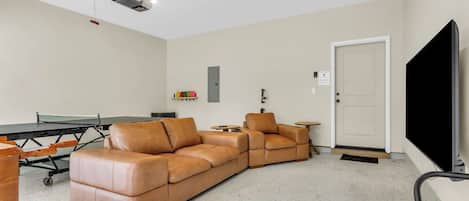 A living room with a sectional sofa, a ping pong table, a flat-screen TV, and a projector mounted on the ceiling.