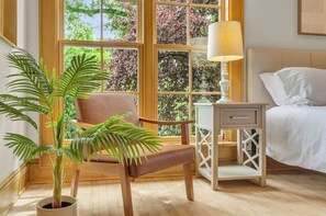 A bedroom with a bed, a bedside table with a lamp, a chair, and a large window with a view of trees.