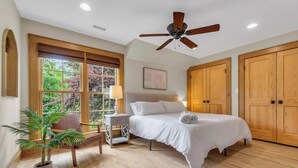 Une chambre à coucher comprenant un lit, un ventilateur de plafond, une fenêtre avec des stores, une chaise, une table de chevet et une plante en pot.