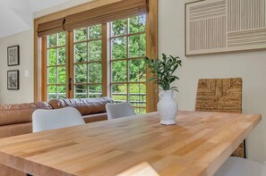 A dining area with a wooden table, white chairs, a brown leather couch, and a large window with a view of greenery.