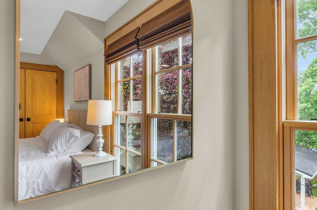 A bedroom with a large window, a bed with white bedding, a bedside table with a lamp, and a wooden wardrobe.