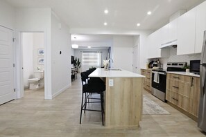 Una cocina moderna con una isla central, taburetes negros y un comedor con mesa y sillas blancas.