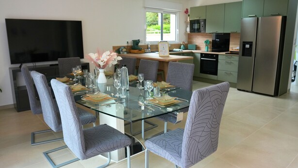 A modern kitchen with a dining area, featuring a glass table, grey upholstered chairs, and stainless steel appliances.