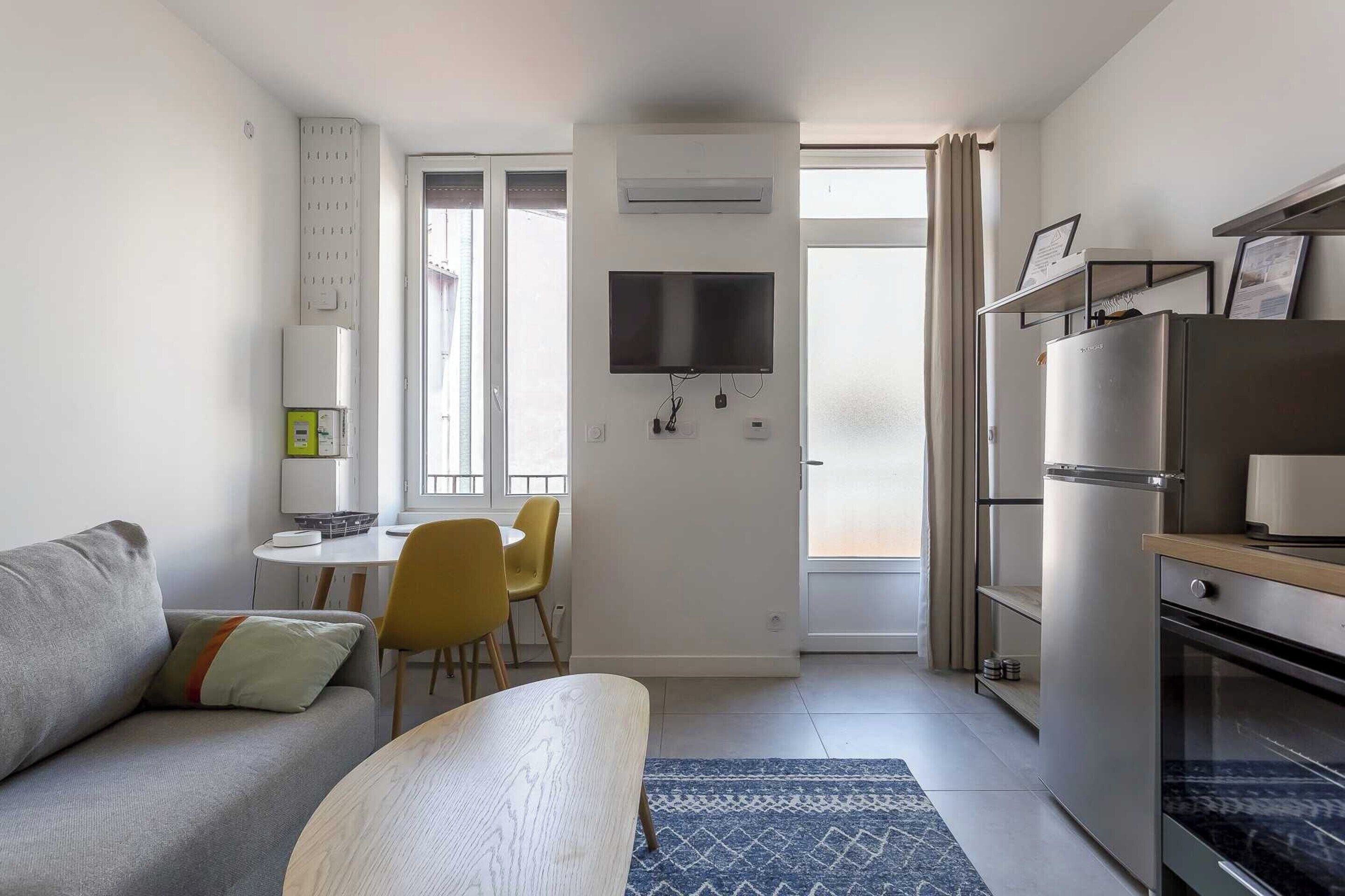 A compact living space with a sofa, a dining table with chairs, a kitchen area with a refrigerator, and a flat-screen TV mounted on the wall.