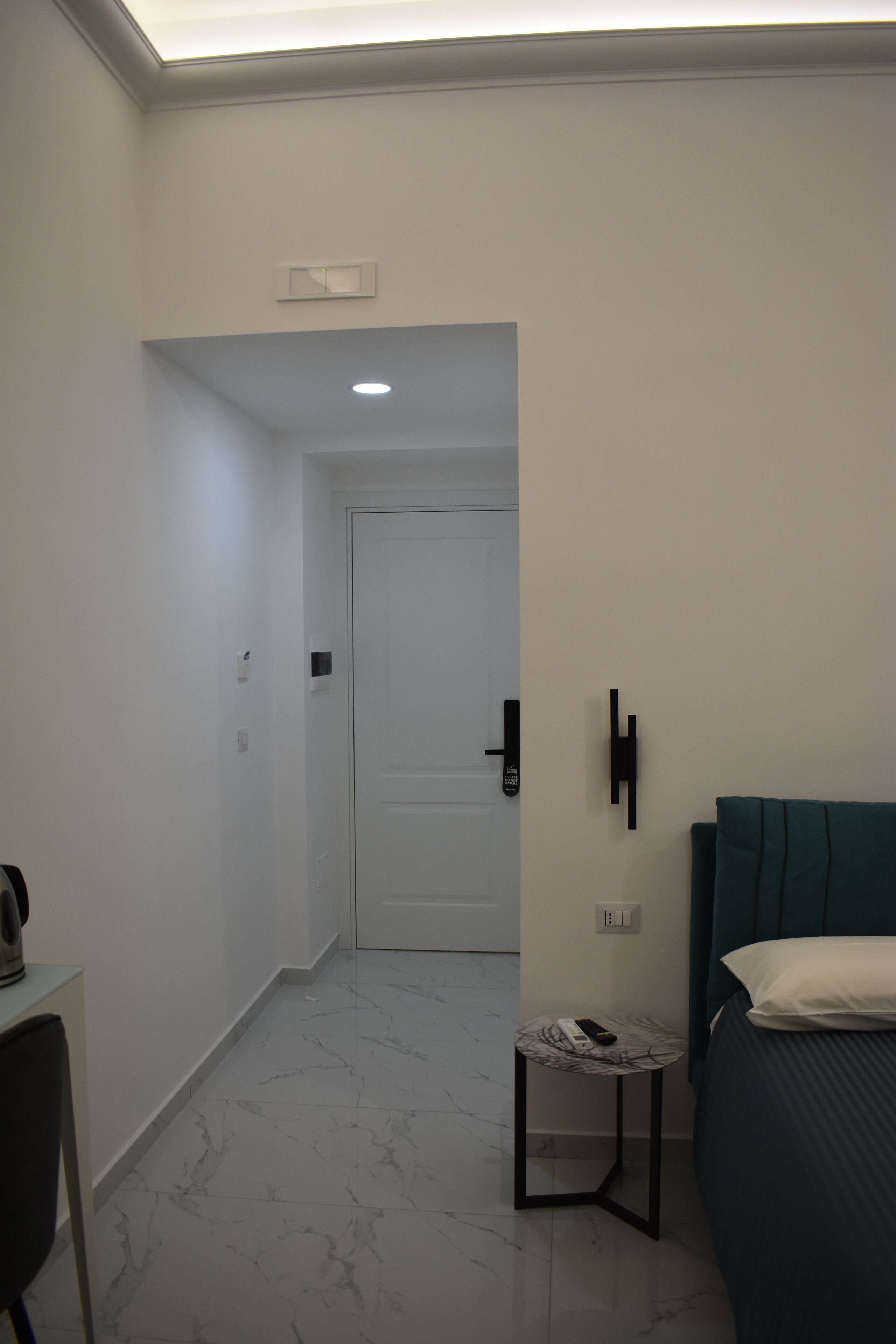 A hotel room with a bed, a small table with a remote control, a chair, and a door leading to another room.