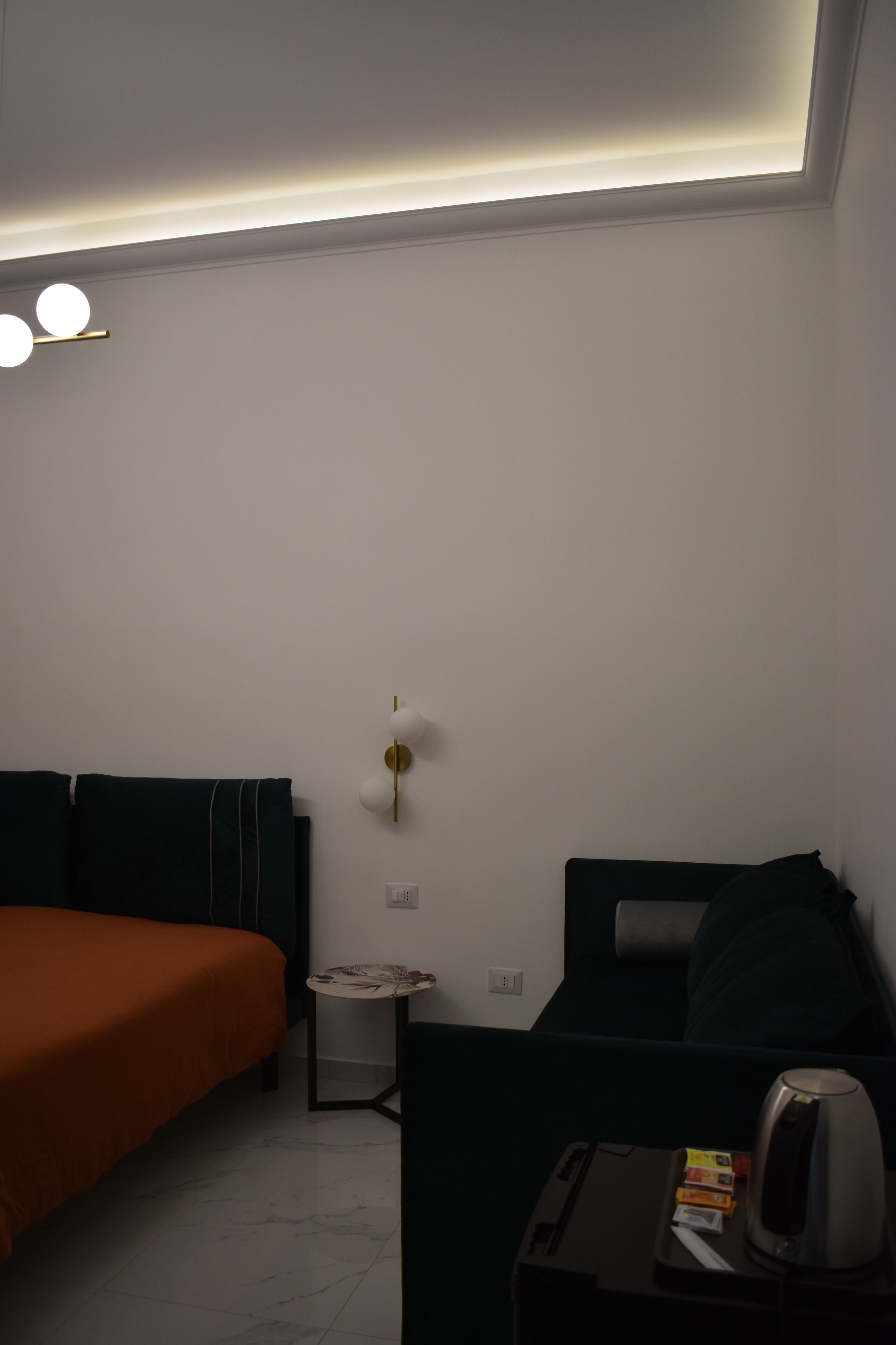 Habitación de hotel con una cama, una mesita con hervidor y una lámpara fijada a la pared.