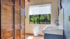 Una cocina con armarios de madera, una ventana con vistas a la vegetación, un cubo de basura blanco y un lavamanos empotrado.