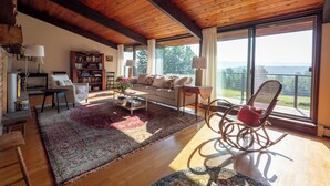 Un vaste salon doté d’un plafond en bois, de grandes fenêtres offrant une vue sur l’extérieur, un coin salon confortable avec un canapé et des fauteuils, et une bibliothèque remplie de livres.