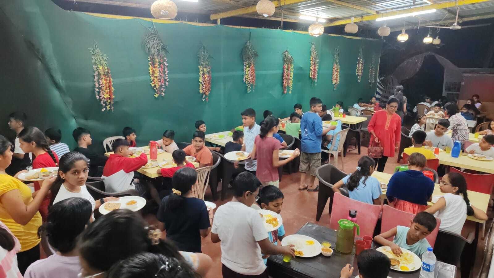 A crowded dining area with tables and chairs, people eating, and hanging decorations on the wall.