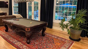 A pool table with a decorative chandelier, a large window with curtains, and a potted plant.