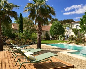 A poolside area with lounge chairs and a swimming pool, surrounded by palm trees and a building with a tiled roof.