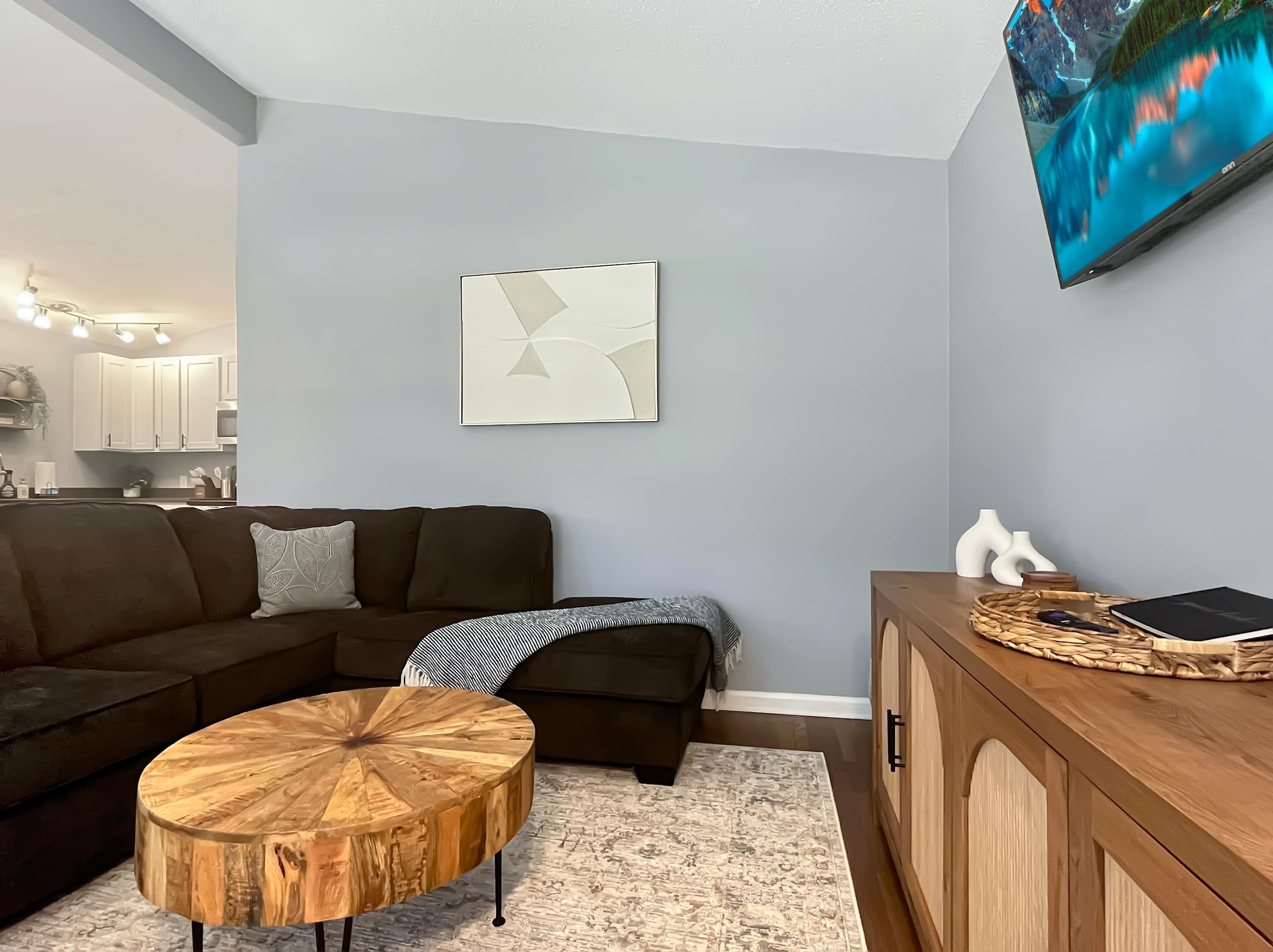 Una sala moderna con un sofá modular, una mesa redonda de madera, una mesita auxiliar y un televisor montado en la pared.