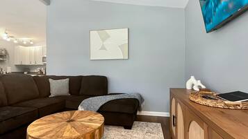 Una sala moderna con un sofá modular, una mesa redonda de madera, una mesita auxiliar y un televisor montado en la pared.