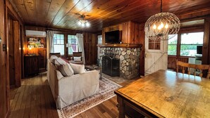 A cozy cabin interior with a stone fireplace, wooden ceiling, and a dining area.
