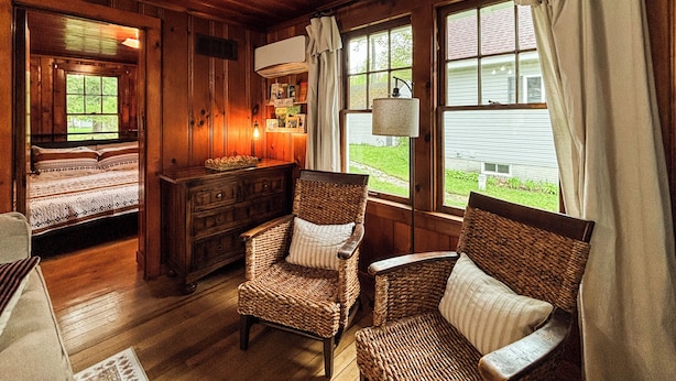 A cozy room with wooden walls, a bed, two wicker chairs, a dresser, and large windows with curtains.