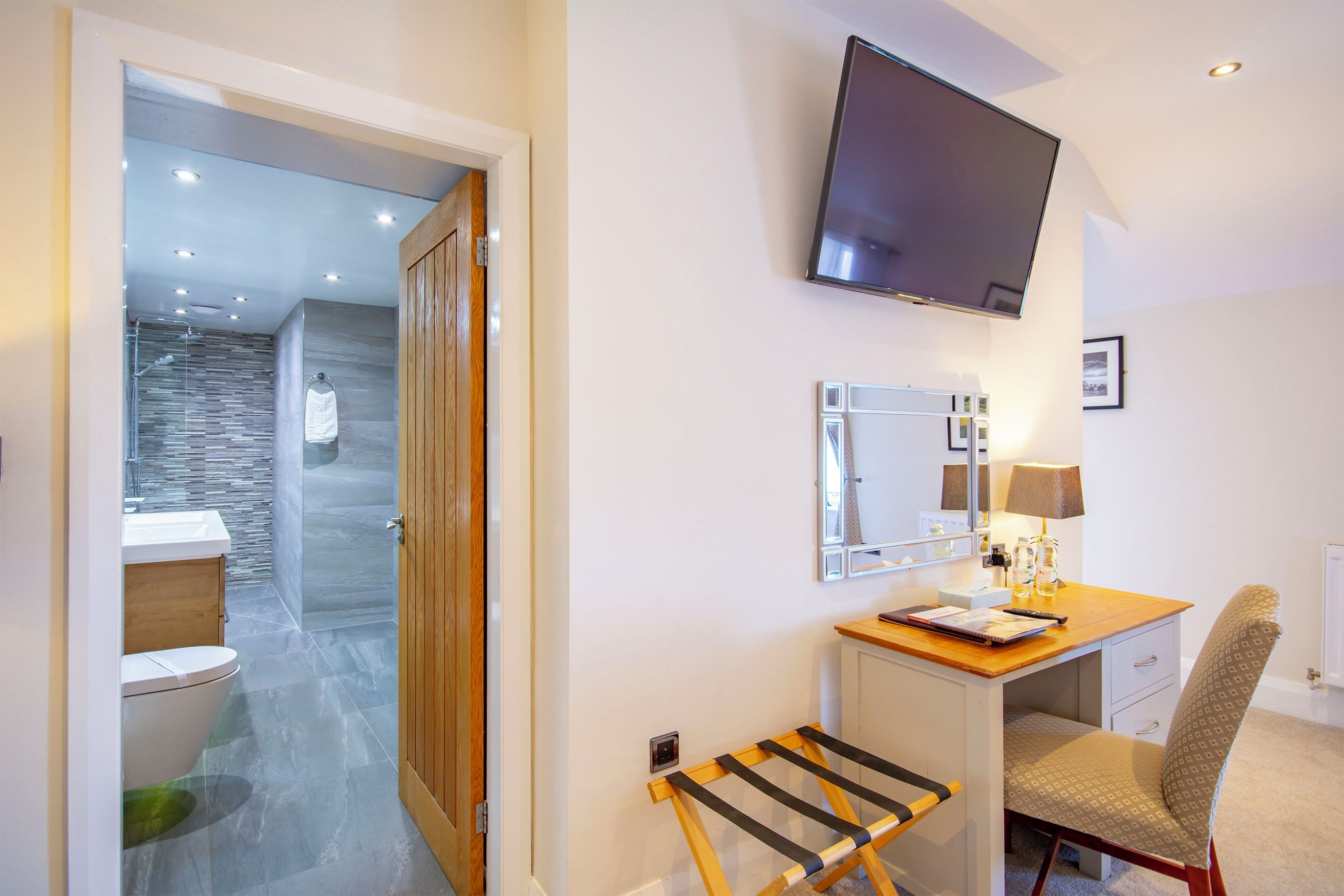 Een hotelkamer met een houten bureau, een stoel, een aan de muur bevestigde televisie, een spiegel en een badkamer die door een geopende deur zichtbaar is.