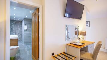 Una habitación de hotel con un escritorio de madera, una silla, un televisor montado en la pared, un espejo y un baño visible a través de una puerta abierta.