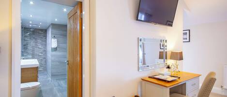 A hotel room with a wooden desk, a chair, a television mounted on the wall, a mirror, and a bathroom visible through an open door.