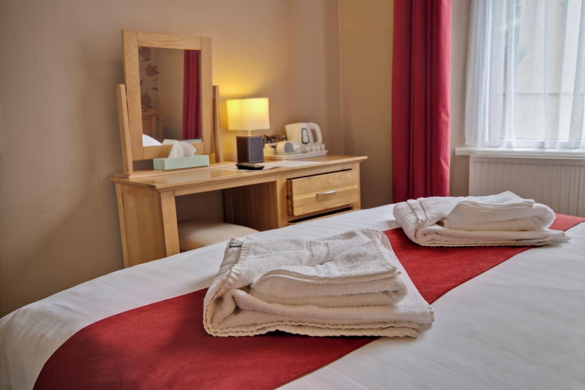 A hotel room with a bed, a wooden desk, a mirror, a lamp, and a window with curtains.
