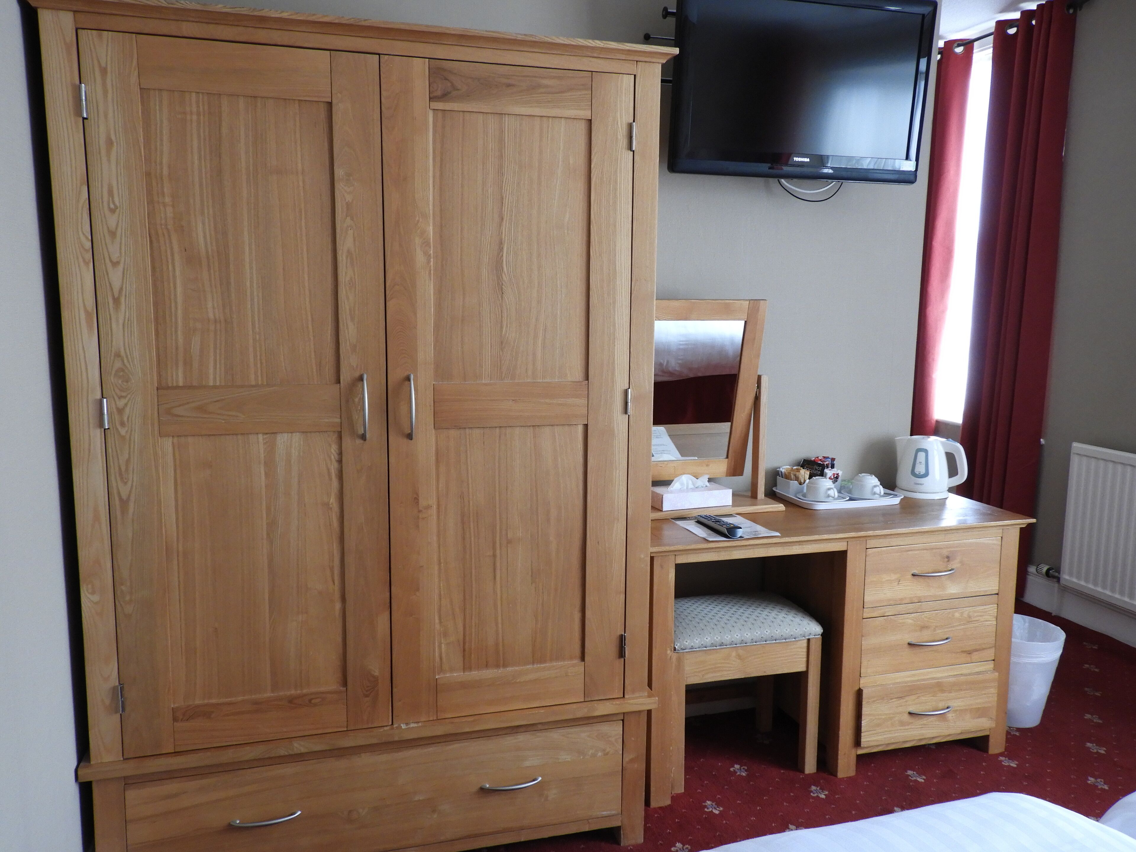 Une armoire en bois avec des tiroirs, un miroir, un bureau avec une chaise, une télévision et un radiateur.
