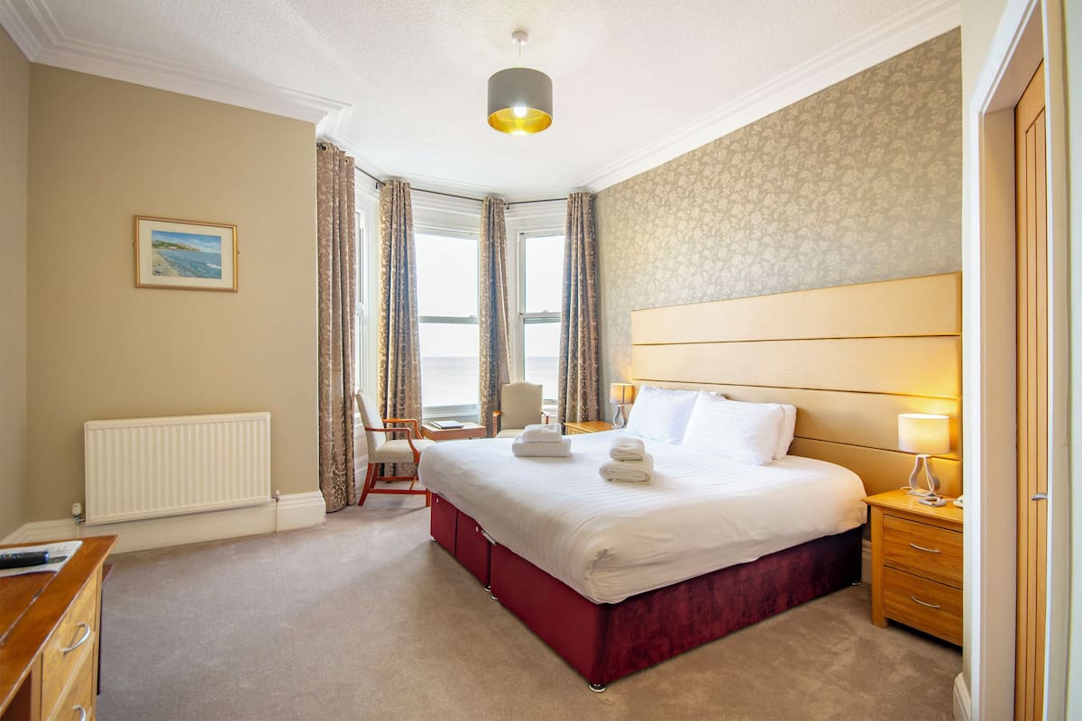 A hotel room with a large bed, a desk, a chair, a radiator, and a window with curtains.