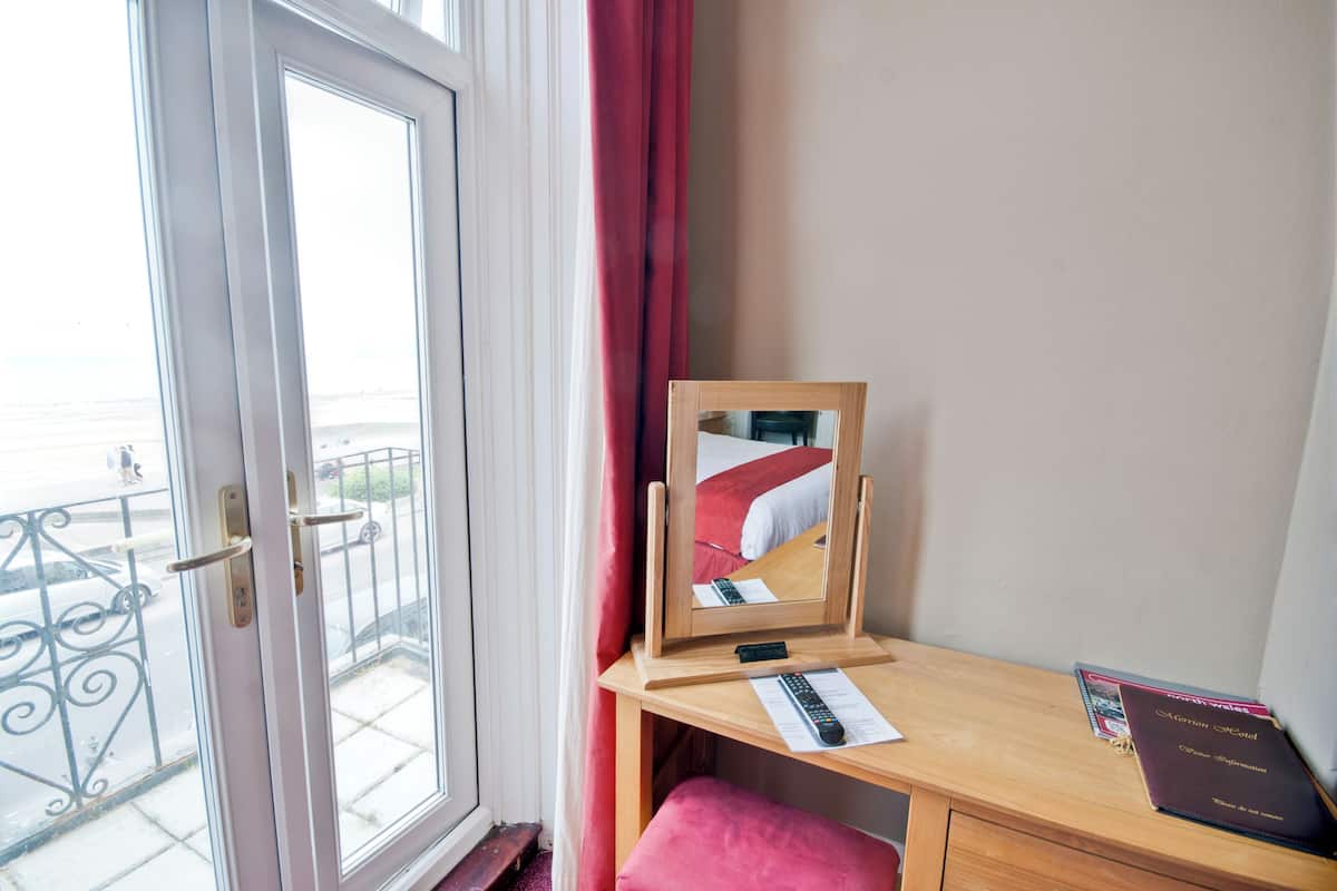 A hotel room with a wooden desk, a mirror, a red curtain, a window with a view of the street, and a remote control on the desk.