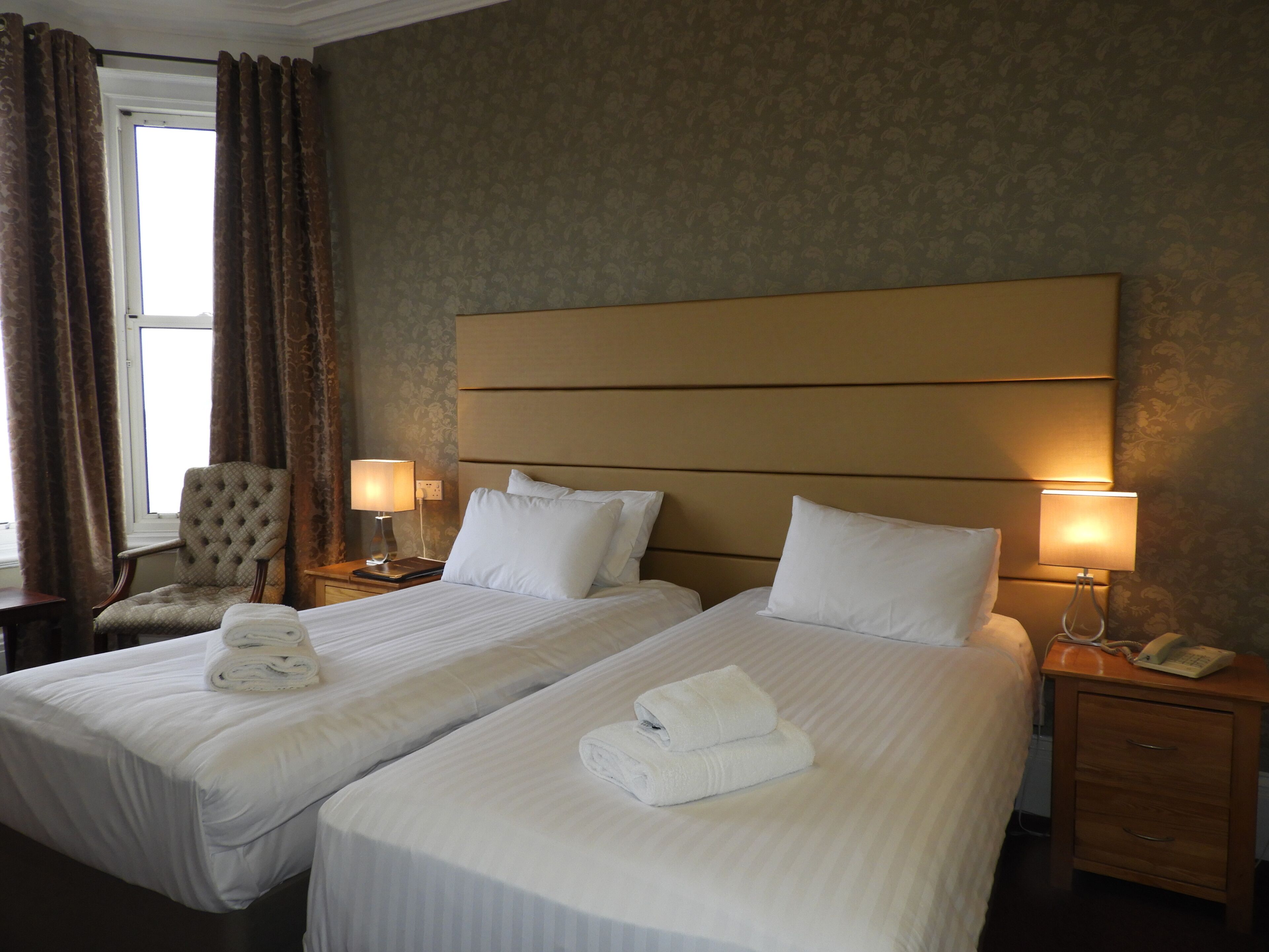A hotel room with two beds, a chair, lamps, a telephone, and a window with curtains.