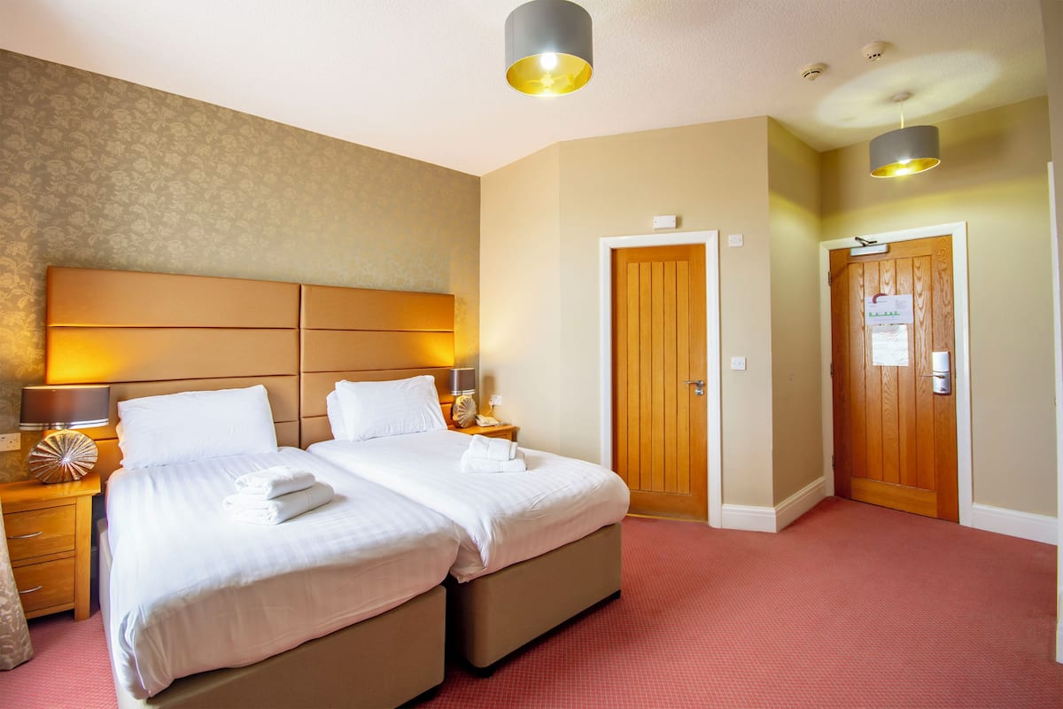 A hotel room with a double bed, two bedside tables, a nightstand, a wooden door, and a red carpeted floor.