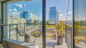 A balcony with two chairs, a small table, and a potted plant, overlooking a cityscape with modern buildings and a clear sky.