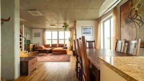 Un salón amplio con un sofá grande, mesa de comedor y sillas. Tiene suelo de madera, una alfombra y un ventanal que proporciona luz natural.