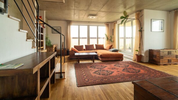 Un salón amplio con suelo de madera, un sofá marrón, una mesa de centro y una escalera con barandilla.