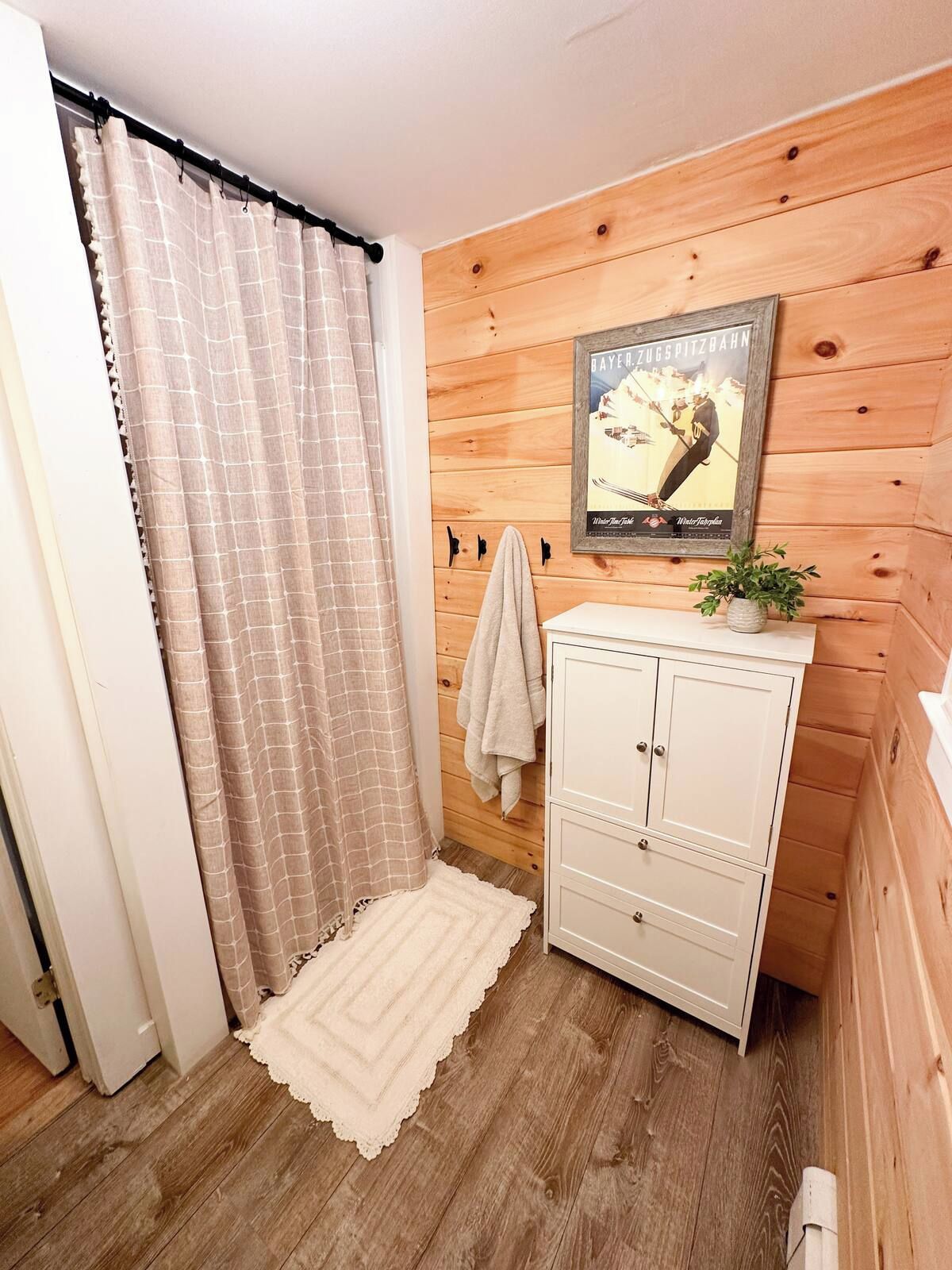 Ett badrum med träpanel på väggarna, ett vitt skåp, en inramad tavla, en handduksställning och en mönstrad gardin.