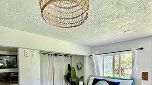 Une chambre avec une couvre-lit turquoise, un luminaire suspendu en fibres tressées, une vue sur la verdure à l’extérieur et une porte blanche donnant sur une salle de bain.