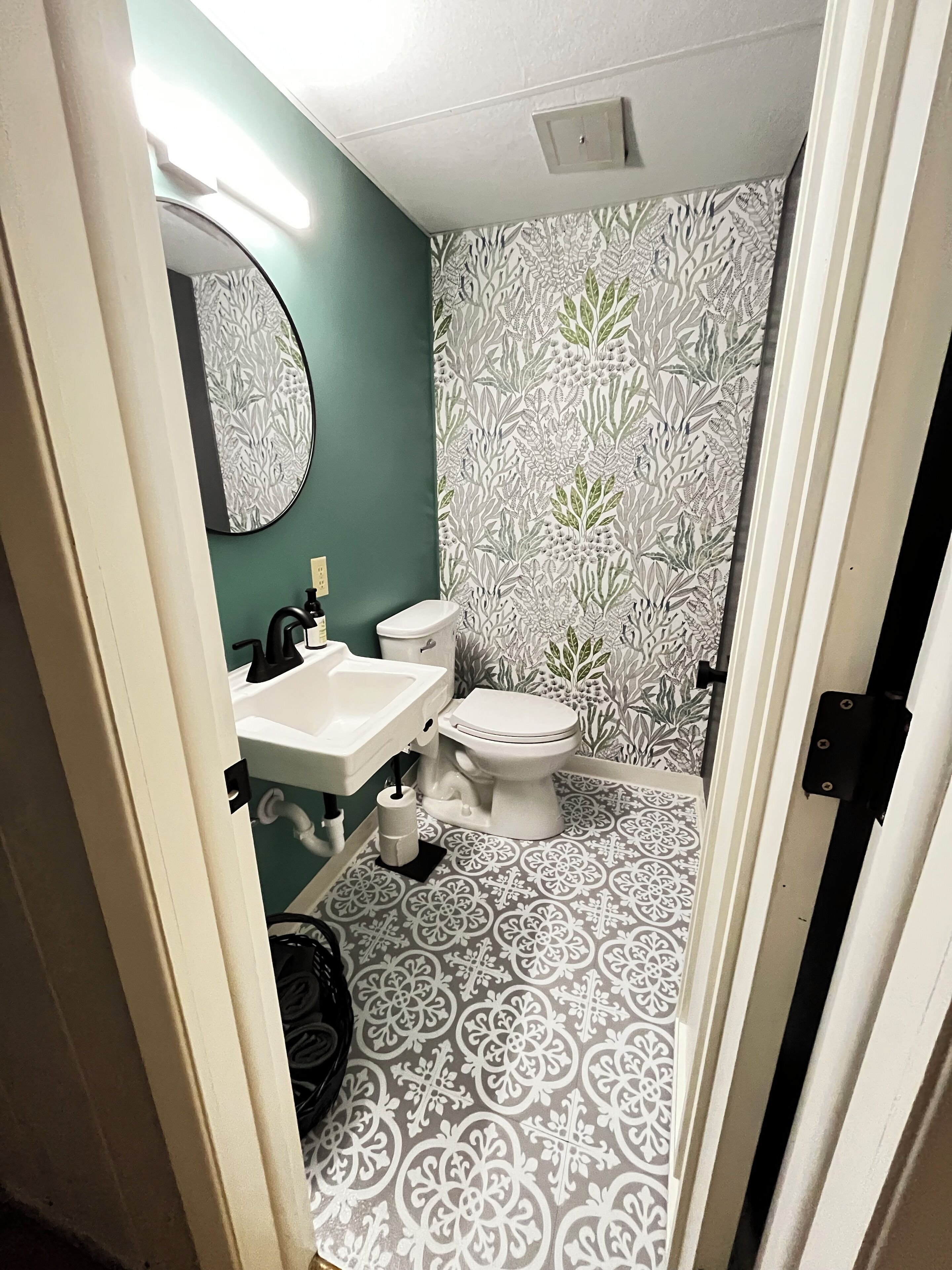 Une salle de bain comprenant des toilettes, un lavabo et un miroir. Les murs sont décorés d’un papier peint à motifs.