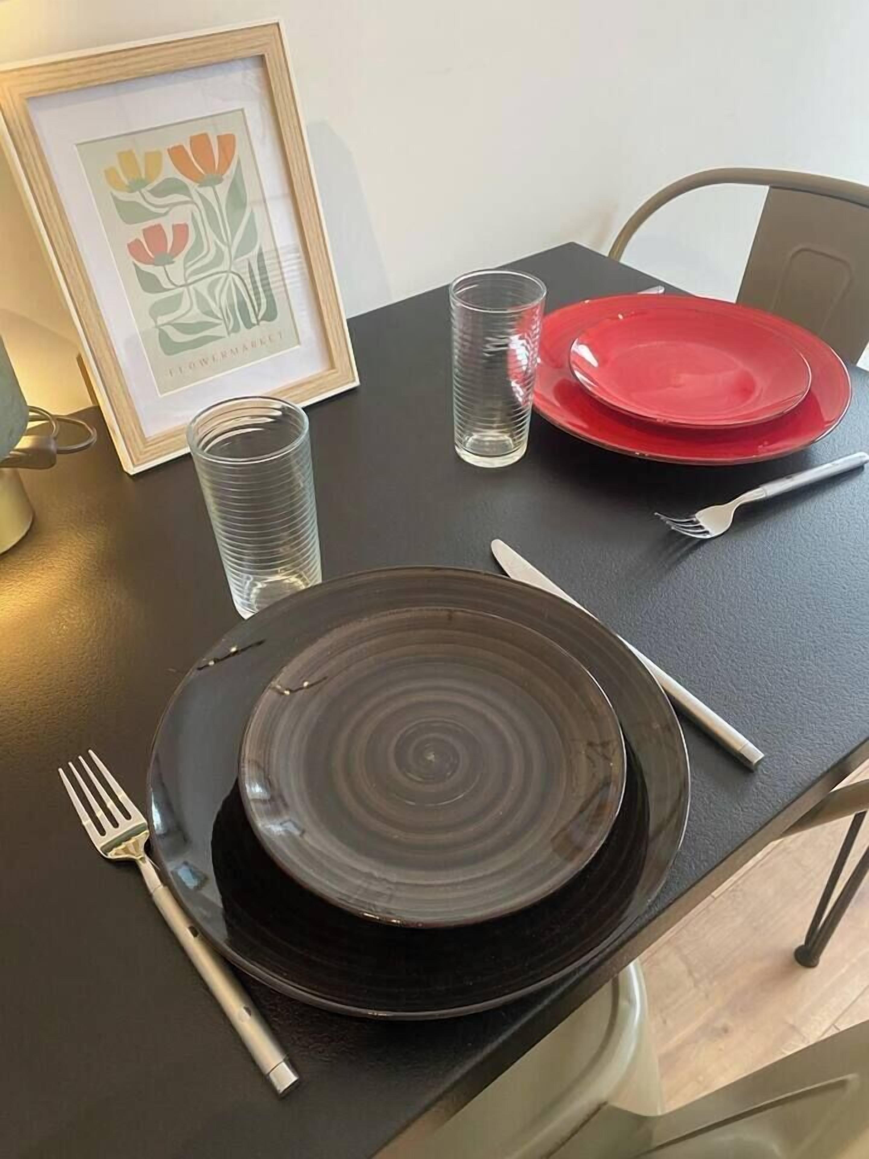 A table set with a black plate, a red plate, a clear glass, a fork, a knife, and a framed picture.
