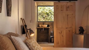 A bedroom with a bed, pillows, a nightstand, a wooden wardrobe, a window with a view of greenery, and a small kitchenette with a sink and a refrigerator. - New years special! Your private jungle escape! (Quepos)