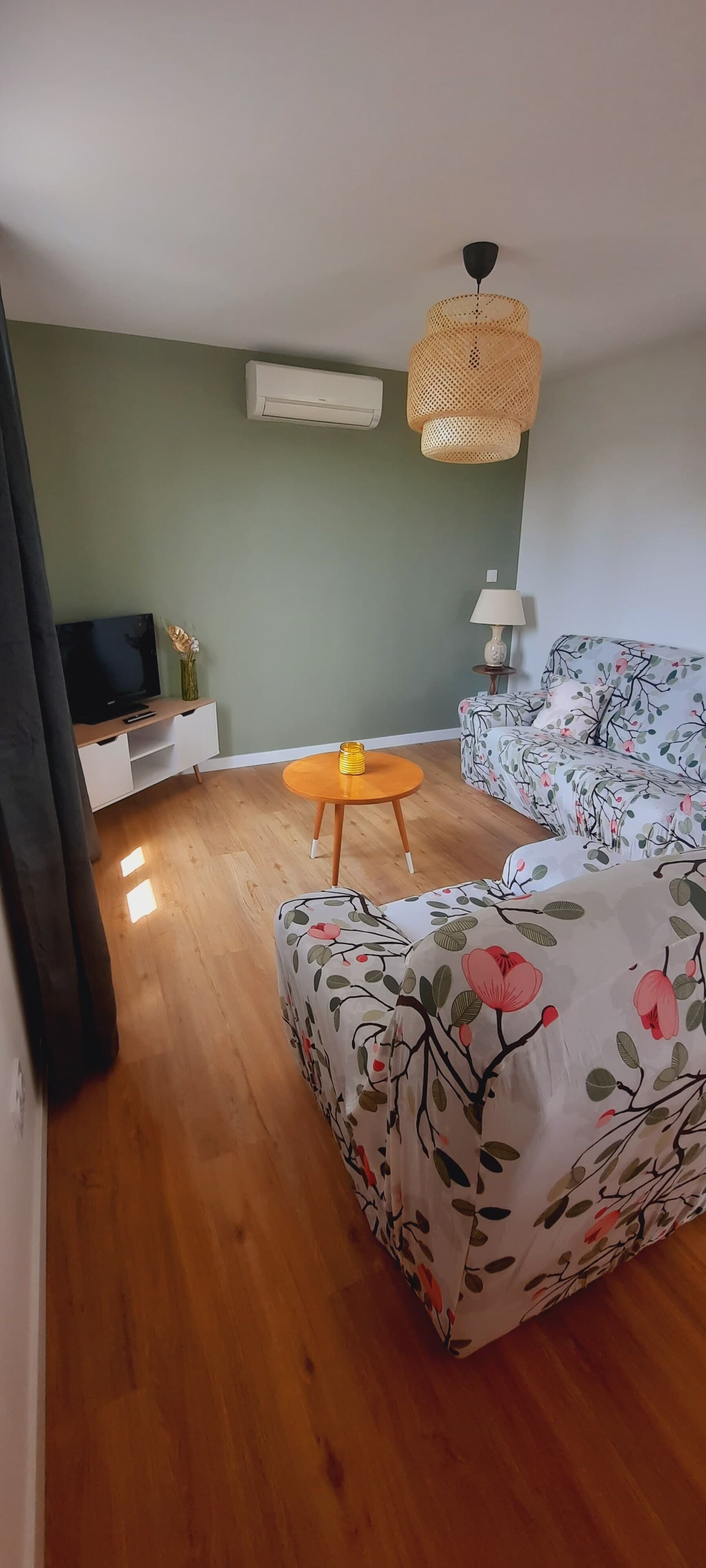 A room with a wooden floor, a sofa with floral upholstery, a small round table with a lamp, and a television on a white cabinet.