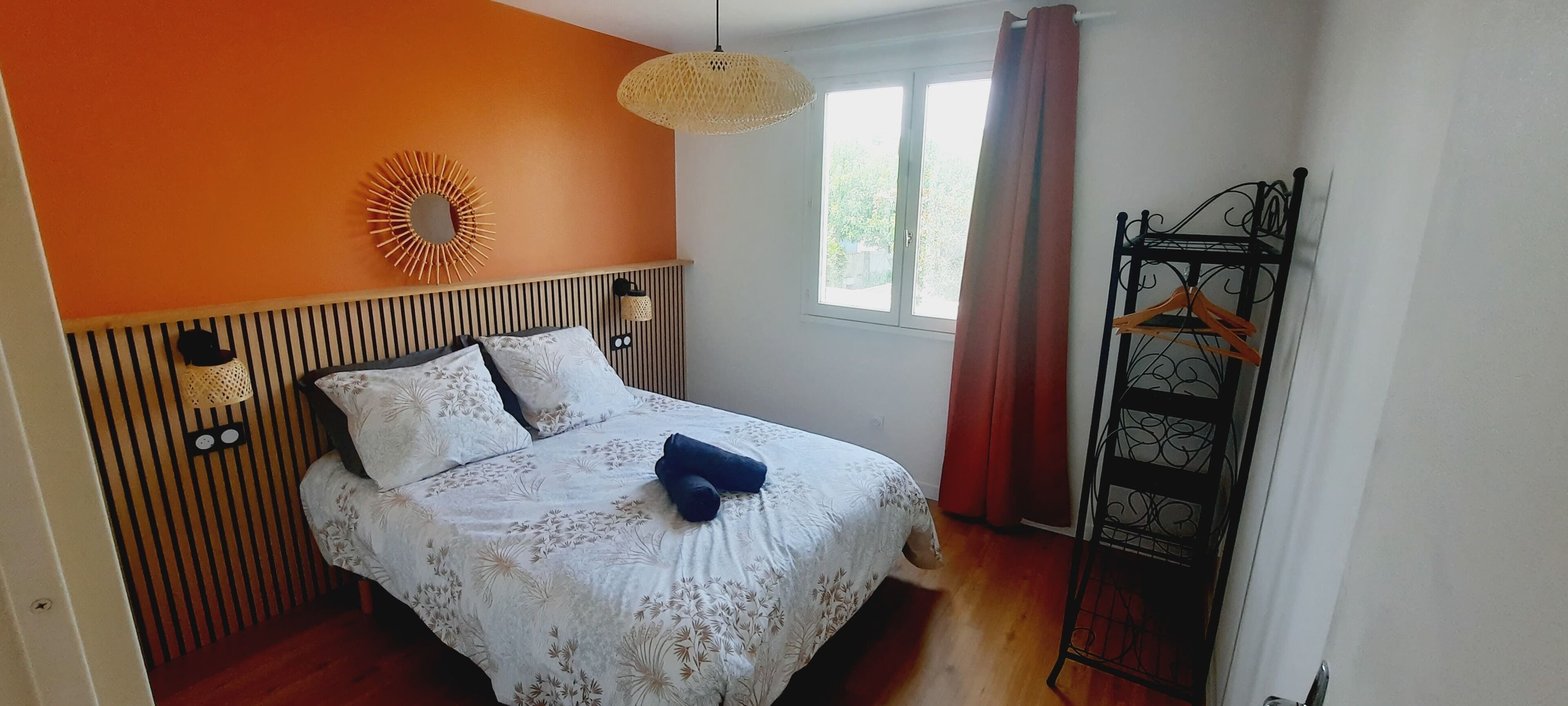 Une chambre à coucher avec un lit, un luminaire suspendu, une fenêtre avec des rideaux et un portant métallique avec des vêtements.