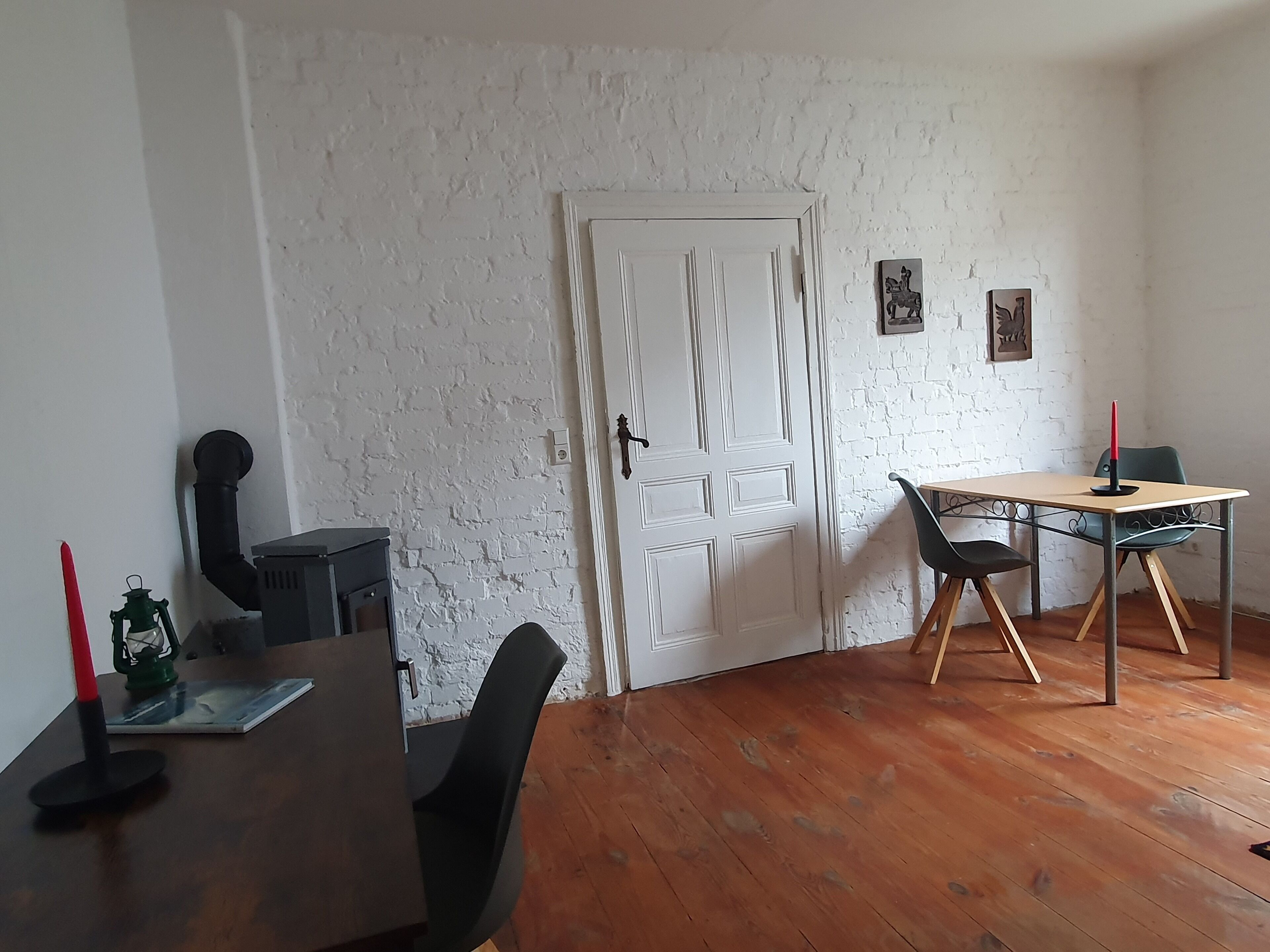 A room with a white brick wall, a wooden door, a table with chairs, and a small shelf with a green lantern and a red candle.