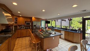 A modern kitchen with wooden cabinets, a central island, bar stools, and a large window offering a view of greenery.