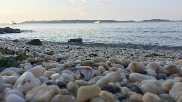 A pebbled beach with calm water, distant boats, and a clear sky.